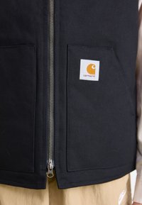 Schwarzes Baumwoll-Tanktop mit zwei vorderen Taschen, silbernem Reißverschluss und einem weißen quadratischen Aufnäher mit orangenem Logo. Strukturierter Stoff, robuste Verarbeitung.
