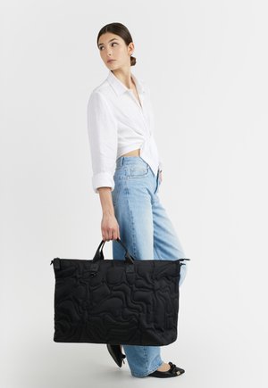 Donna con camicia bianca e jeans azzurri chiaro che tiene una grande borsa da viaggio nera trapuntata per le maniglie.