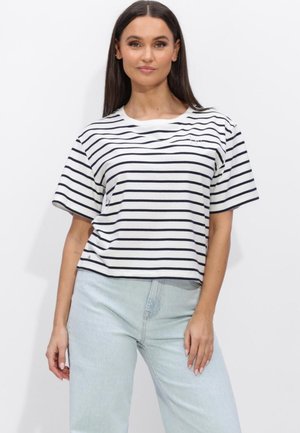Vrouw met lang donker haar draagt een wit-zwart gestreept T-shirt met korte mouwen en lichtblauwe jeans met hoge taille, staand tegen een witte achtergrond.