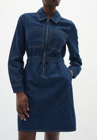 Spijkerjurk met lange mouwen, kraag, twee borstzakken en een rits aan de voorkant. Beschikt over een elastische taille en een donkerblauwe kleur met zichtbare stiksels.