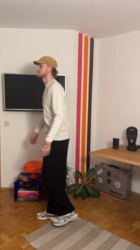 Καθημερινή εμφάνιση με ένα ελαφρύ πουλόβερ, μαύρο παντελόνι και ασημένια αθλητικά παπούτσια. Το φόντο περιλαμβάνει μια τηλεόραση, πολύχρωμες ρίγες στον τοίχο και εσωτερικά φυτά.