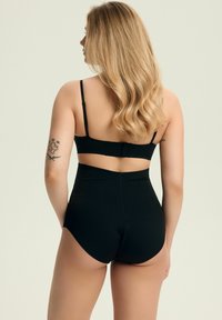 Blondynka z falującymi włosami, ubrana w czarne majtki wysoki stan i cz bra, stojąca tyłem z tatuażami na ramionach.