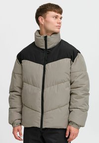 Gewatteerde jas in beige en zwart. Heeft een hoge kraag, ritssluiting en chevronstiksels. Gemaakt van een glad, waterafstotend materiaal.