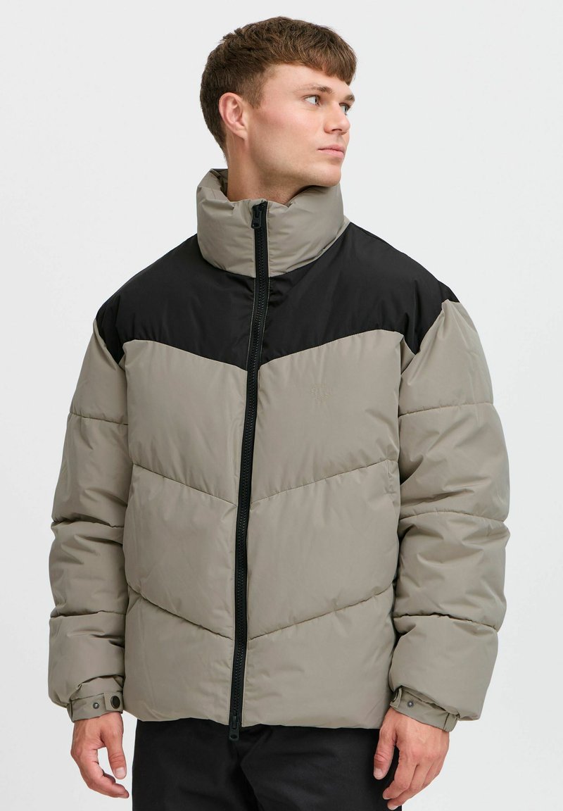Gewatteerde jas in beige en zwart. Heeft een hoge kraag, ritssluiting en chevronstiksels. Gemaakt van een glad, waterafstotend materiaal.
