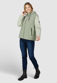 Veste en polaire verte et doudoune beige avec capuche, poches zippées noires et surface texturée ; associée à un jean sombre et des bottes à lacets noires.