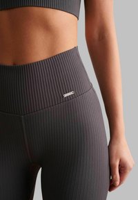 Leggings grigi a coste con vita alta, texture liscia e piccolo logo in metallo accentato sulla cintura.
