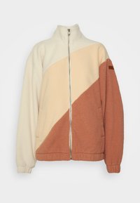 Chaqueta de forro polar con cuello alto, que presenta un diseño de bloques de color en beige, crema y terracota, con cremallera completa y bolsillos laterales.