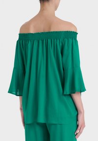 Top verde con spalle scoperte, scollatura arricciata, maniche fluttuanti e tessuto leggero. Presenta una vestibilità ampia e una texture liscia.