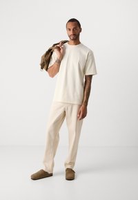 Uomo con tatuaggi indossa una maglietta crema e pantaloni, scarpe slip-on marroni, tiene una giacca marrone sulla spalla davanti a uno sfondo neutro.