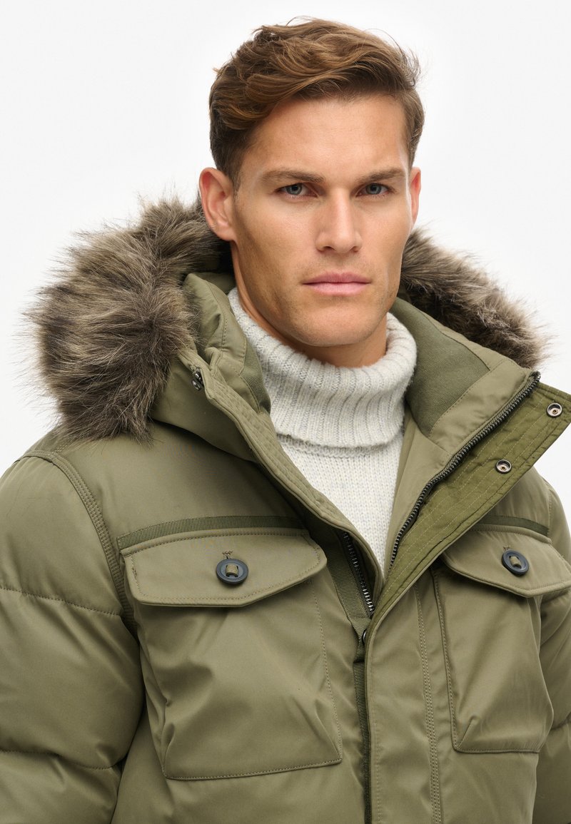 Chaqueta acolchada verde oliva con capucha de piel sintética desmontable, múltiples bolsillos frontales y cierre con cremallera, llevada sobre un suéter gris claro.