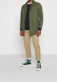 Man som bär en olivgrön jacka, svart tröja, beige byxor, vita strumpor och gröna högstövlar som står på en grått golv.