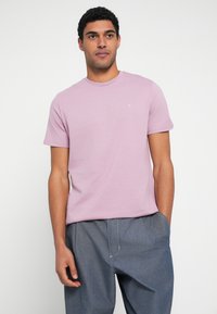 Šviesiai rožinė medvilninė marškinėliai su apvalia kaklo linija ir trumpomis rankovėmis, krūtinėje mažas baltas logotipas. Derinama su pilkomis kelnėmis.