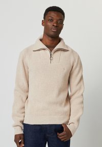 Pull en tricot côtelé beige avec un col zippé à quart, présentant un tissu texturé et un petit logo sur la poitrine. Porté avec un jean foncé.