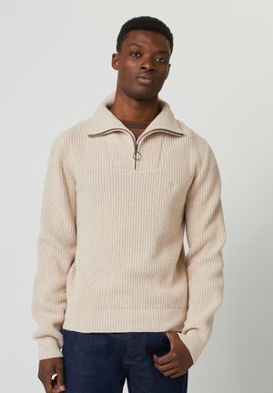 Pull en tricot côtelé beige avec un col zippé à quart, présentant un tissu texturé et un petit logo sur la poitrine. Porté avec un jean foncé.