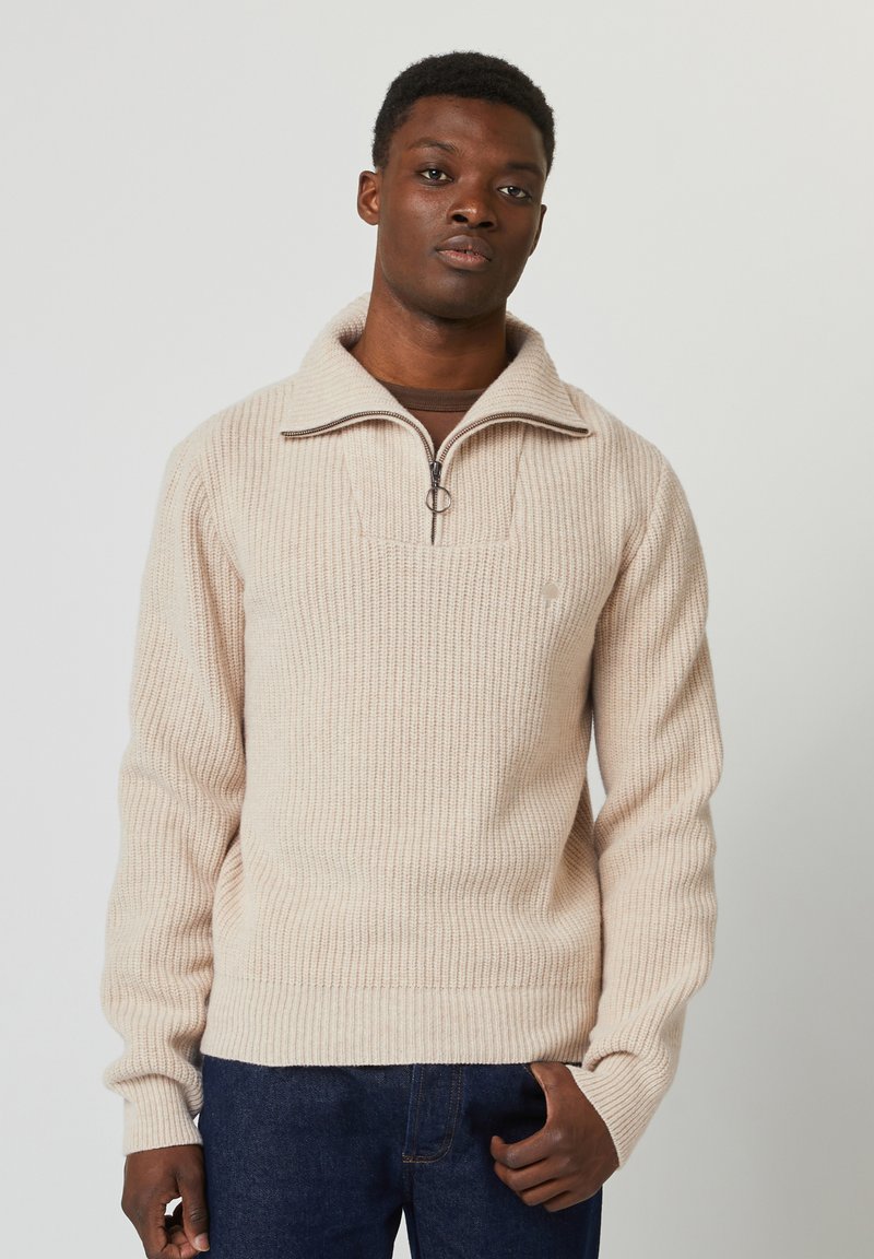 Pull en tricot côtelé beige avec un col zippé à quart, présentant un tissu texturé et un petit logo sur la poitrine. Porté avec un jean foncé.