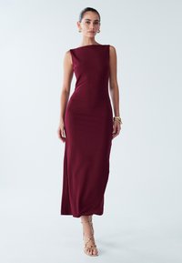 Burgundy sleeveless maxi dress with a boat neckline, body-hugging fit, side slit, and smooth fabric texture, paired with strappy gold sandals.
