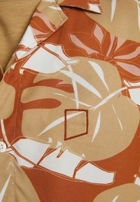 Camicia a maniche corte, con bottoni, in tessuto beige con grandi motivi di foglie arancioni e marroni. Presenta una toppa quadrata bordata con cuciture marroni.