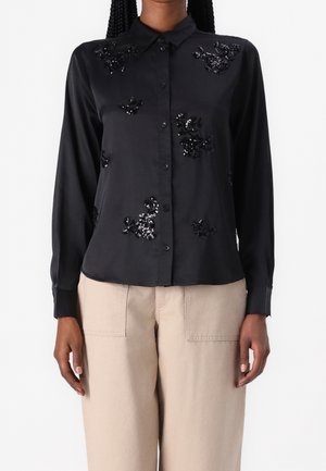 Schwarzes langärmeliges Hemd mit Knöpfen, verziert mit glänzenden schwarzen Pailletten in Blumenform, kombiniert mit beigen hoch taillierten Hosen mit aufgesetzten Taschen vorne.