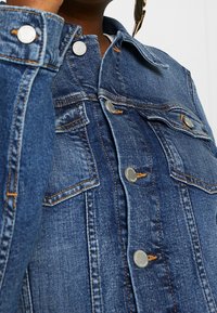 Denimjacka med en medelblå nyans, silverdetaljer, knäppningar, bröstfickor och kontrasterande orange sömmar.