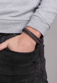 Bruin gevlochten leren armband, ronde vorm, gedragen om de pols. Grijze trui zichtbaar, hand rustend in een zwarte denimzak.