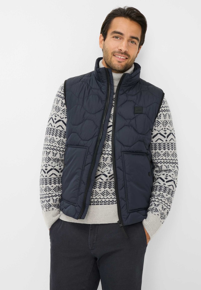 Marineblauer gesteppter Weste mit hohem Kragen, Reißverschluss vorne und zwei Taschen, über einem gemusterten cremefarbenen Pullover und dunklen Hosen getragen.