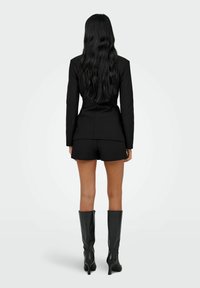 Zwarte blazer met gestructureerde schouders, gecombineerd met zwarte shorts. Model draagt kniehoge, glanzende zwarte laarzen. Het haar is lang en golvend.