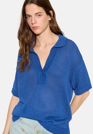 Młoda kobieta z długimi brązowymi włosami, ubrana w luźną niebieską dzianinową koszulkę polo z krótkim rękawem i jasne spodnie z kwiatową haftem.