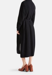 Černé šaty z vlněné směsi s pásem, dlouhými rukávy a bočními rozparky; doplněné hnědými botami na blokovém podpatku z lesklé kůže.