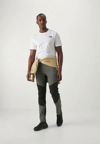 Men's outfit featuring a white T-shirt, gray and black pants with different textures, and a beige sweater tied at the waist. Black shoes.