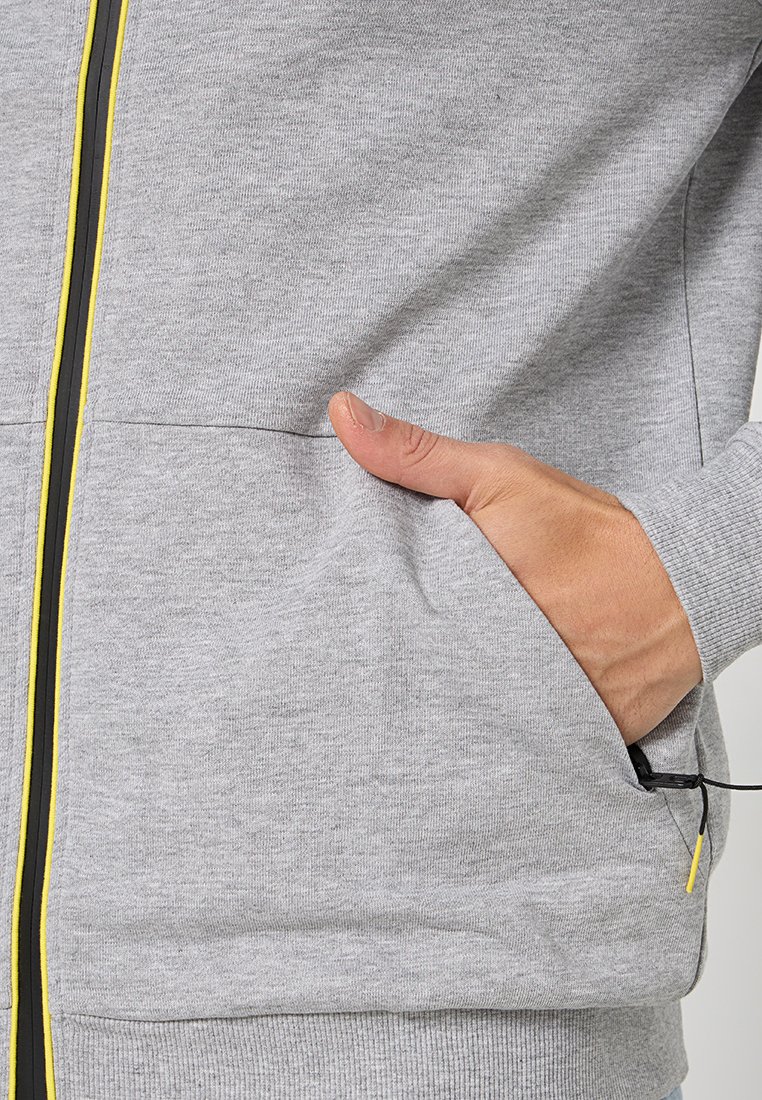 Sweatshirt gris avec fermeture éclair sur le devant, zips accent jaunes et gris, et une poche latérale. Tissu doux et texturé avec des poignets côtelés.