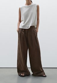 Haut sans manches gris clair associé à un pantalon en lin marron ample. Le pantalon présente des plis avant et des poches latérales, porté avec des sandales noires.