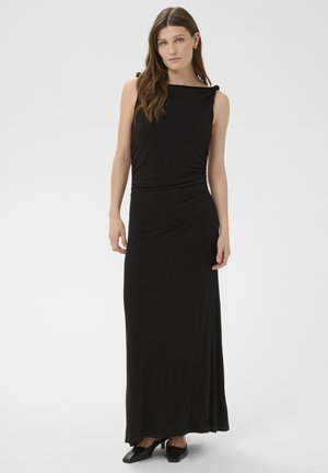 Femeie în picioare, purtând o rochie maxi neagră fără mâneci, cu detalii pliate pe partea laterală și pantofi negri cu vârf ascuțit, pe un fundal simplu, deschis la culoare.