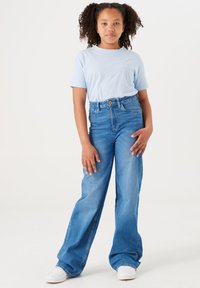 Camiseta de color azul claro, jeans de tiro alto y pierna ancha de color azul con tejido texturizado. Zapatillas blancas con perforaciones y diseño de cordones.