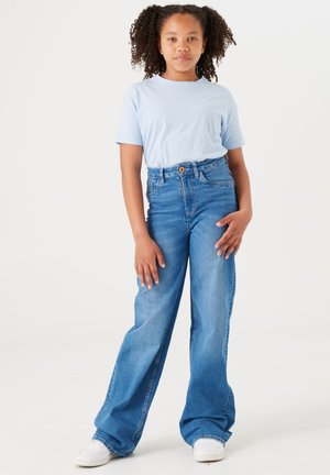 Hellblaues T-Shirt, hochgeschnittene, weit geschnittene Jeans in Blau mit strukturiertem Stoff. Weiße Sneakers mit Perforationen und Schnürdesign.