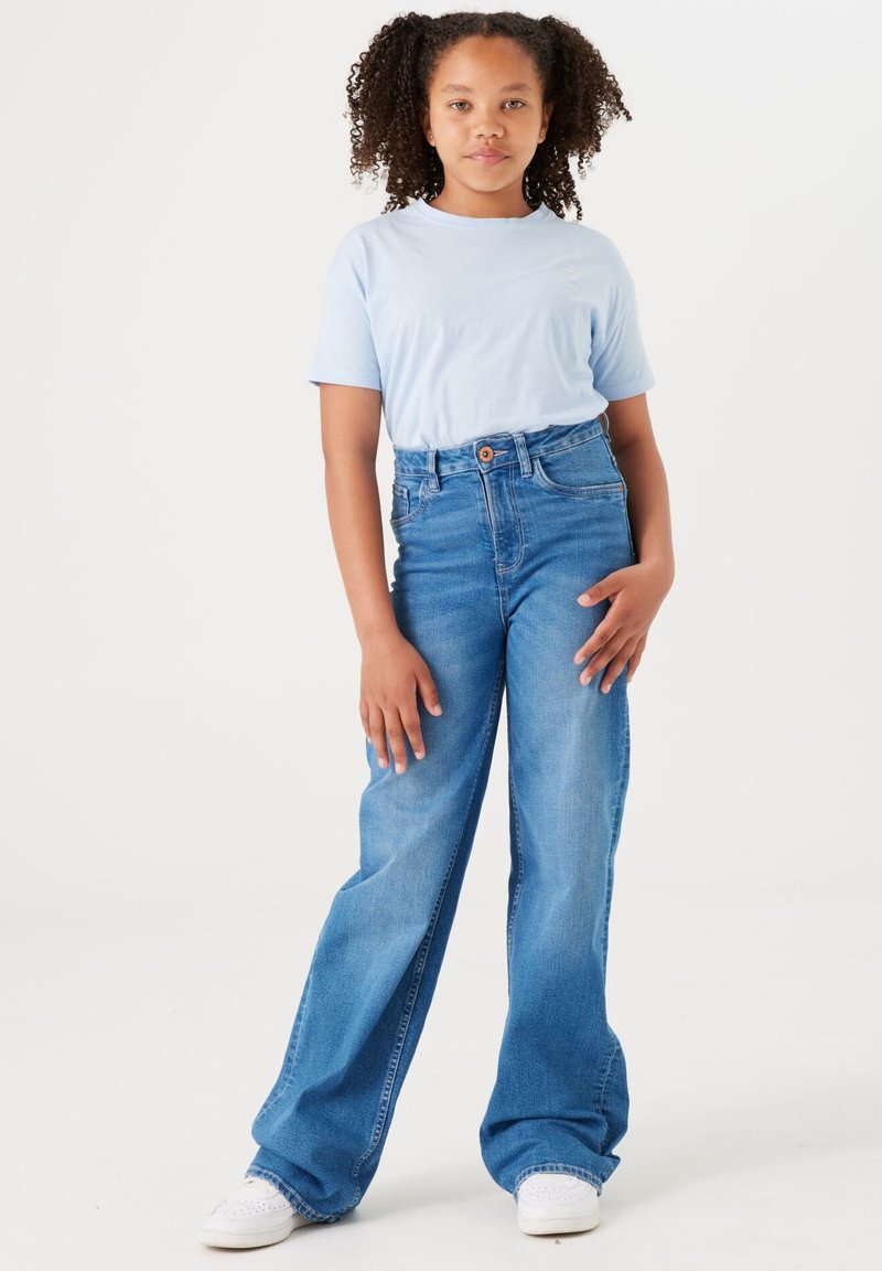 Camiseta de color azul claro, jeans de tiro alto y pierna ancha de color azul con tejido texturizado. Zapatillas blancas con perforaciones y diseño de cordones.