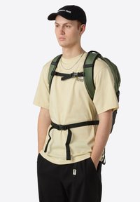Green backpack with multiple straps, black hardware, worn by a person in a beige shirt and black pants, standing against a white background.