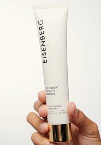 Tube blanche avec du texte noir et doré. L'étiquette indique "EISENBERG" et "EXFOLIANT LISSANT EXPRESS." Accent doré à la base, tenu par une main.
