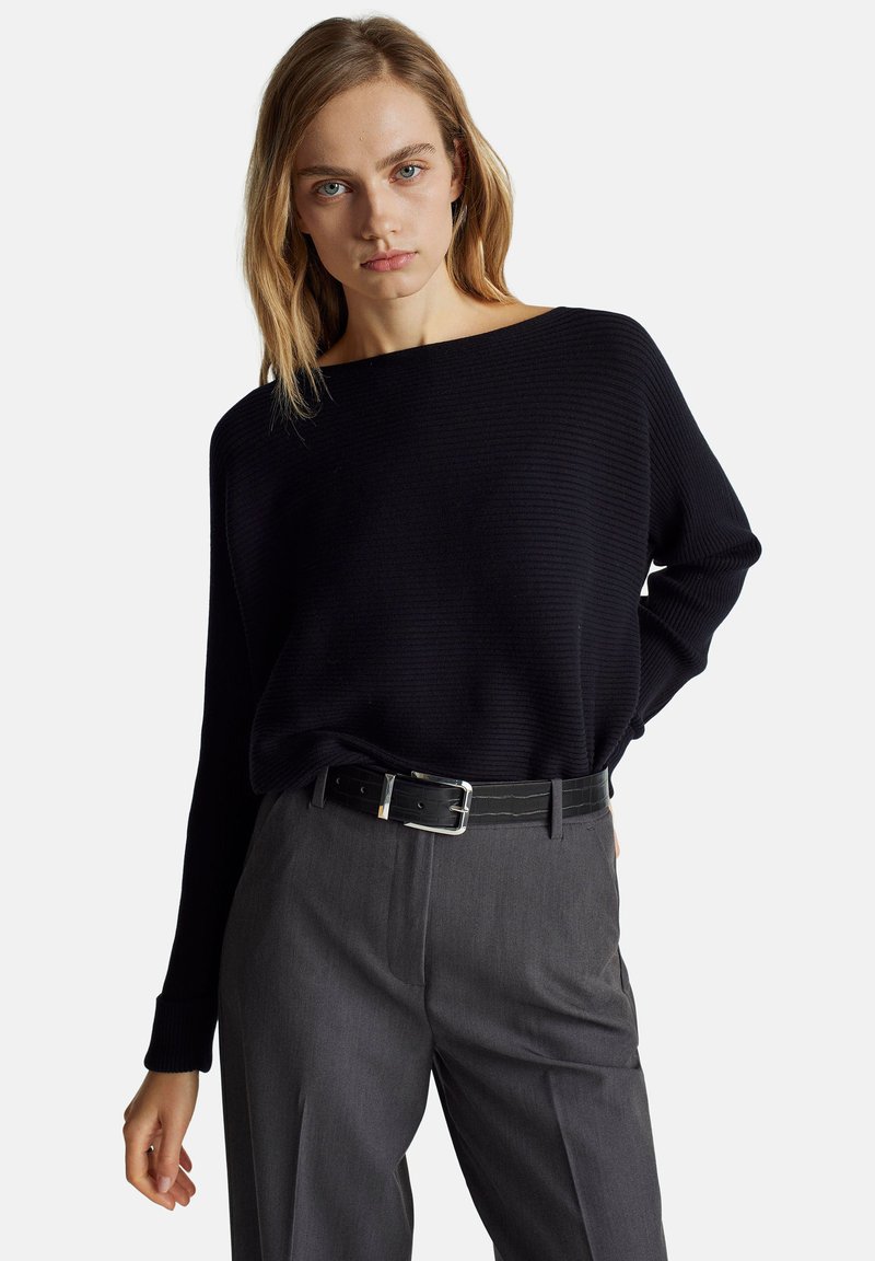 Czarny sweter prążkowany o szerokim dekolcie, noszony na szarych spodniach z wysokim stanem, podkreślony czarnym paskiem z srebrną klamrą.