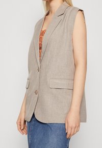 Beige ärmelloser Blazer aus strukturiertem Stoff, mit Reverskragen, zwei vorderen Taschen und Knopfverschluss, getragen über einem gemusterten Oberteil.