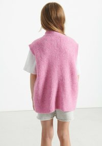 Roza pletena brezrokavna jopica z rebrasto teksturo, oblečena čez belo srajco, v paru z sivimi kratkimi hlačami z razfrcanimi robovi.