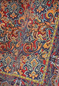 Tkanina z misternymi wzorami florystycznymi i paisley w odcieniach pomarańczowym, niebieskim i zielonym na czerwonym tle, wykończona żółtymi akcentami.