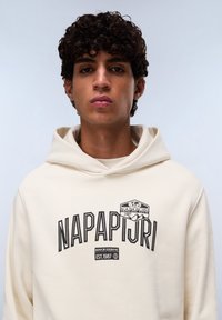 Beige hoodie met een zwart bedrukte logo dat "NAPAPIJRI 1987" op de borst zegt. Gladde stofstructuur met een ontspannen pasvorm.