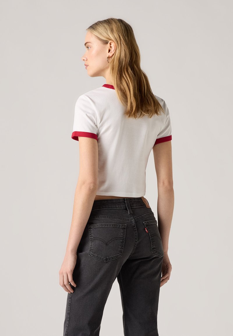T-shirt blanche raccourcie avec des bordures rouges, associée à un jean noir avec poches arrière et étiquette en cuir, présentée de côté.