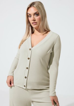 Blonde Frau trägt eine beige gerippte, geknöpfte Strickjacke und passende gerippte Hosen vor einem schlichten hellen Hintergrund.