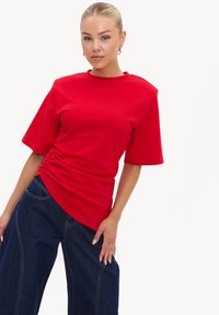 Femme blonde portant un t-shirt rouge à manches courtes froncé et un jean large bleu foncé, posant avec une main sur la hanche.