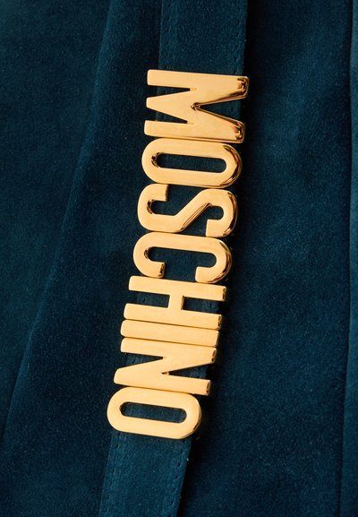 Lettres métalliques dorées formant le mot « MOSCHINO » attachées verticalement sur un tissu bleu foncé, probablement une partie d’un accessoire de mode ou d’un vêtement.