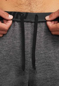 Mains d'un homme ajustant le cordon noir sur un pantalon de sport gris, montrant le bas-ventre avec de légers poils corporels.