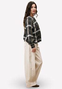 Vrouw met lang golvend haar, gekleed in een zwart-grijs geruite knopshirt en brede beige broek, staande in zijaanzicht met een neutrale uitdrukking.