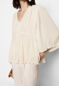 Hellbeige Faltenbluse mit V-Ausschnitt und weiten Ärmeln, mit einem gerafften Bund und weicher Textur. Passt zu hellen Hosen und einer Tasche.