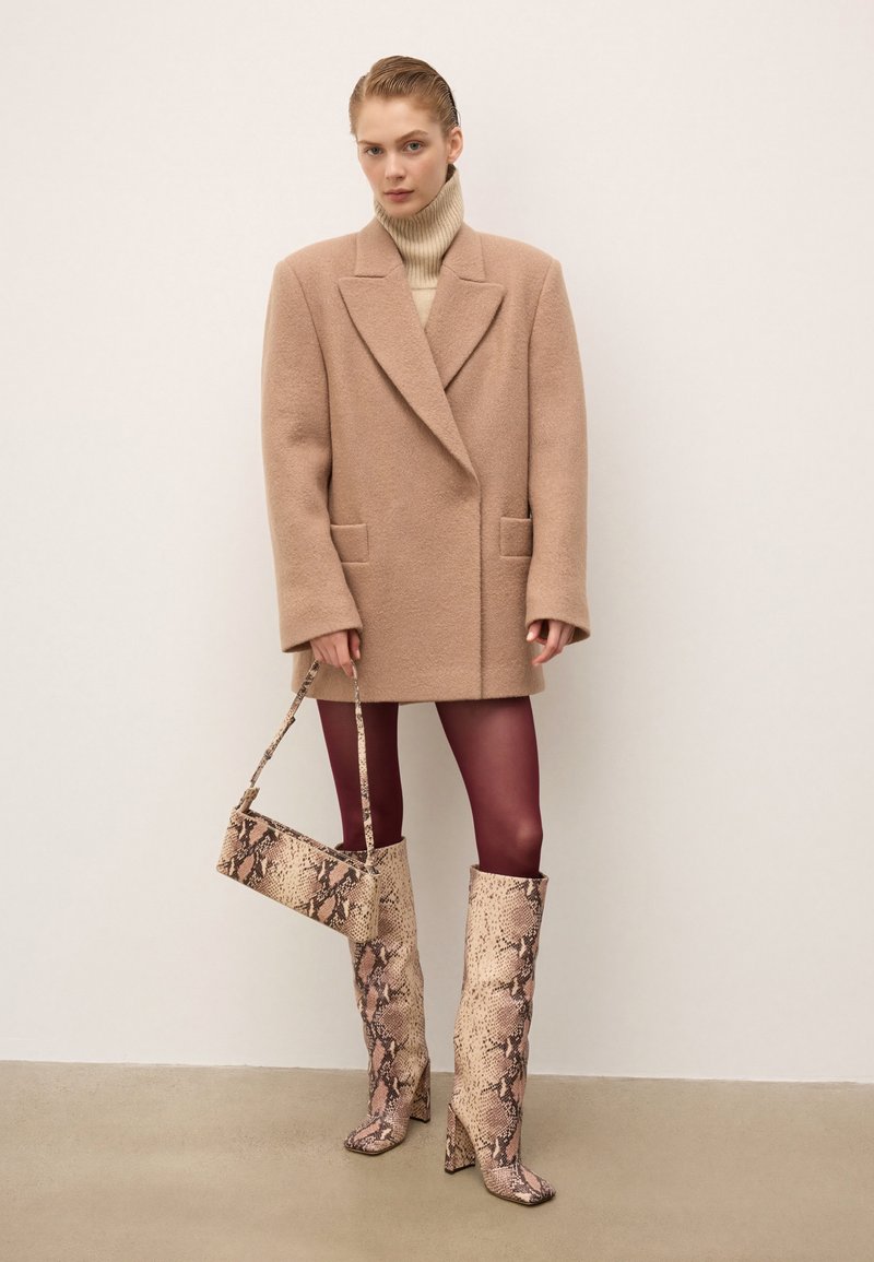 Donna che indossa un cappotto over size beige, un dolcevita crema, collant bordeaux, mentre tiene una borsa in pelle di serpente e stivali fino al ginocchio in pelle di serpente coordinati.